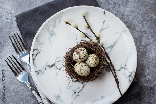 Festive table setting for holiday Easter dinner on gray stone concrete table with copyspace. Quail eggs and willow on plate