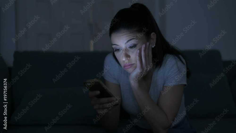 young attractive and tired hispanic 30s woman with mobile phone looking serious and bored using dating app sitting at apartment living room couch late night