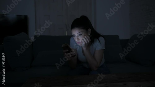 young attractive and tired hispanic 30s woman with mobile phone looking serious and bored using dating app sitting at apartment living room couch late night