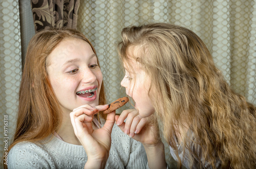 Two sisters, one older teenager with braces smiling and feeding oatmeal cookies to a toddler with long hair