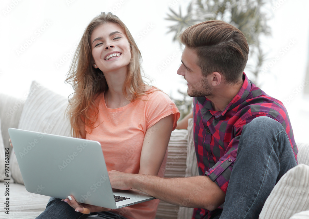 young couple is using a laptop and smiling while sitting on sofa at home