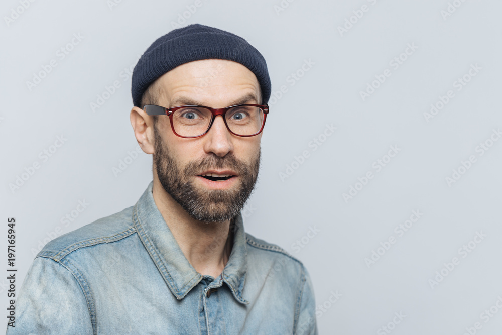 Stylish middle aged male with suprised and curious expression wears spectacles, denim jacket and hat, poses against grey background with copy space for your advertisment or promotional text. Stylish middle aged male with suprised and curious expression wears spectacles, denim jacket and hat, poses against grey background with copy space for your advertisment or promotional text.