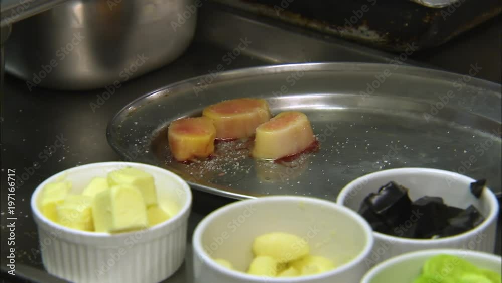 Close up of chef seasoning and then picking up a metal plate with three marinated scallops. Another bowl is filled butter. Close up of chef seasoning and then picking up a metal plate with three marinated scallops. Another bowl is filled butter.