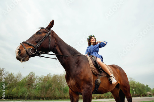 girl with a wreath of flowers on her head sitting in a saddle on a horse on the background of the sky and trees © Ivan