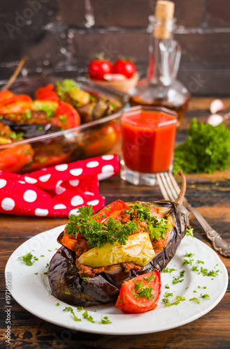 Eggplants stuffed with minced meat and cooked with fresh tomatoes and bell peppers on white plate and in baking dish on wooden rustic table.