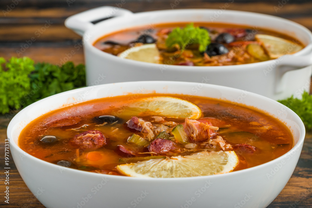Thick and hearty Russian tomato soup Solyanka with turkey, sausages, pickled cucumbers, black olives and fresh lemon in white casserole and bowl on the wooden rustic table. Thick and hearty Russian tomato soup Solyanka with turkey, sausages, pickled cucumbers, black olives and fresh lemon in white casserole and bowl on the wooden rustic table.