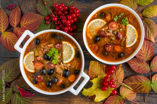Fotografi Thick and hearty Russian tomato soup Solyanka with turkey, sausages, pickled cucumbers, black olives and fresh lemon in white casserole and bowl on the wooden rustic table, top view