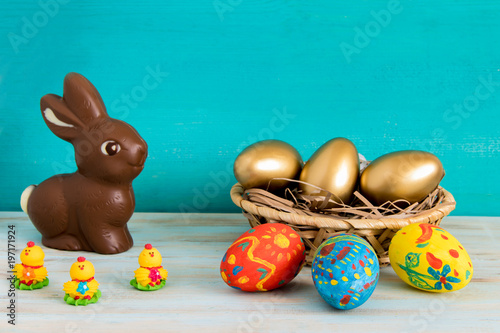 Happy Easter concept with color ang gold aster eggs in basket next to chocolate Easter bunny and baby chicken on blue wooden background photo