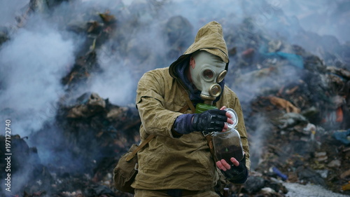 a man in a gas mask holds a sprout in the flask, against the background of fuming debris © Jonny