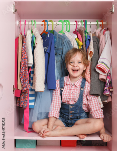 A little girl is sitting in a closet with a children's department. Storage system for children's things