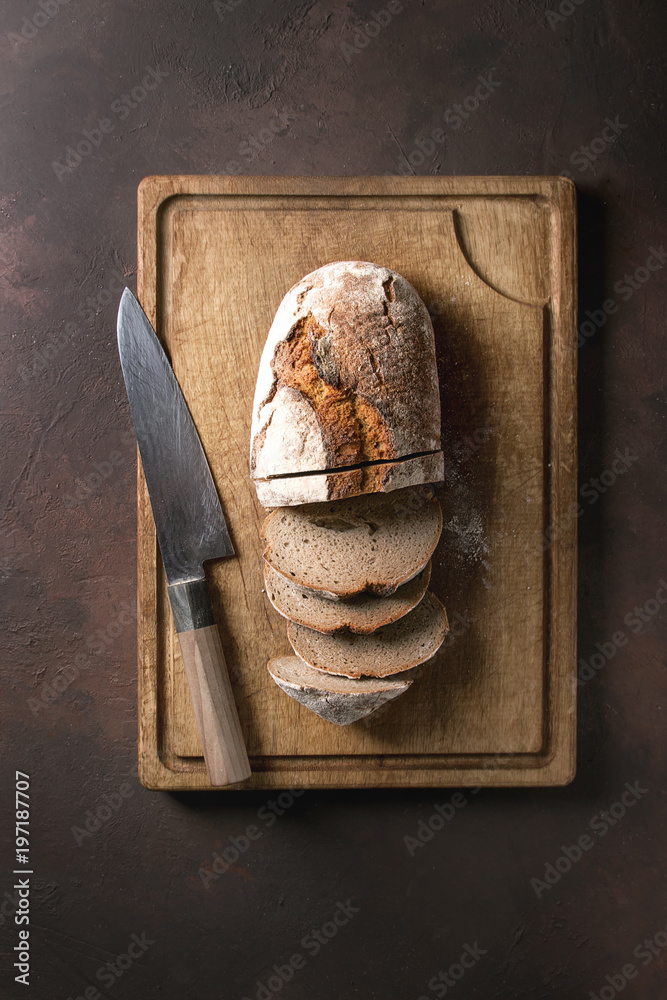 Loaf of fresh sliced baked artisan rye bread on wooden cutting board with knife over dark brown texture background. Top view, copy space. Loaf of fresh sliced baked artisan rye bread on wooden cutting board with knife over dark brown texture background. Top view, copy space.