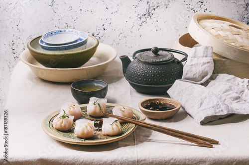 Canvas Print Asian steam potstickers dumplings stuffed by shrimps, served on ceramic plate with soy sesame sauce, chopsticks, teapot, tea, bamboo steamer over kitchen table with linen cloth