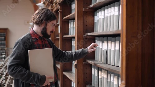 Focused student looking at old archive materials, bearded boy in gray sweater and checked red shirt choosing preferable book, indoor shot in university library