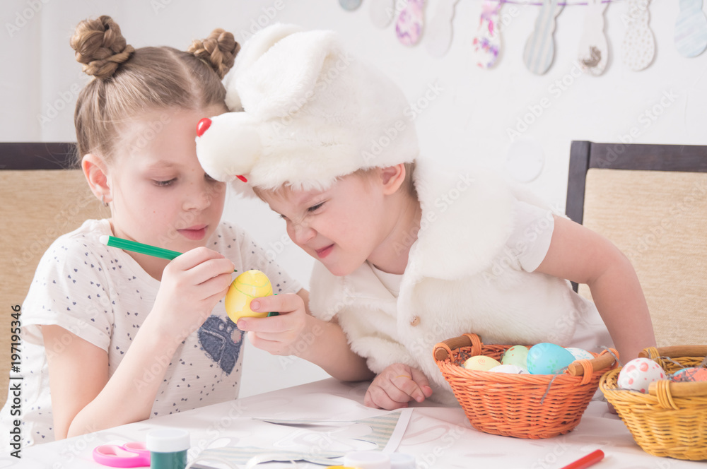 Easter Festival: Children paint Easter eggs at the table. A boy in a rabbit costume looks cunningly at the work of the older sister. Easter Festival: Children paint Easter eggs at the table. A boy in a rabbit costume looks cunningly at the work of the older sister.