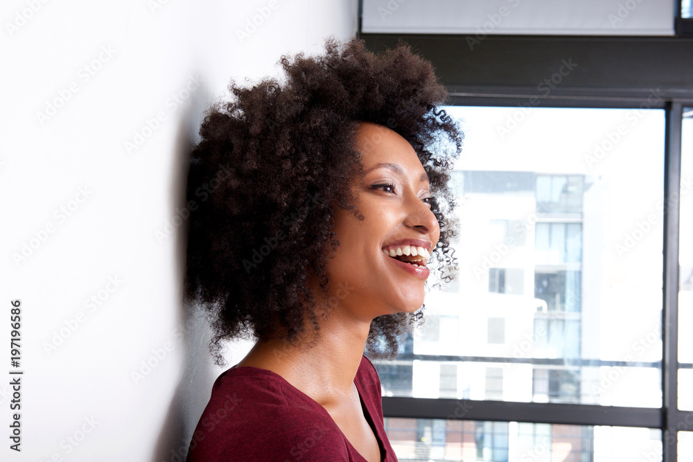 Close up happy young woman with curly hair leaning to wall at home and laughing