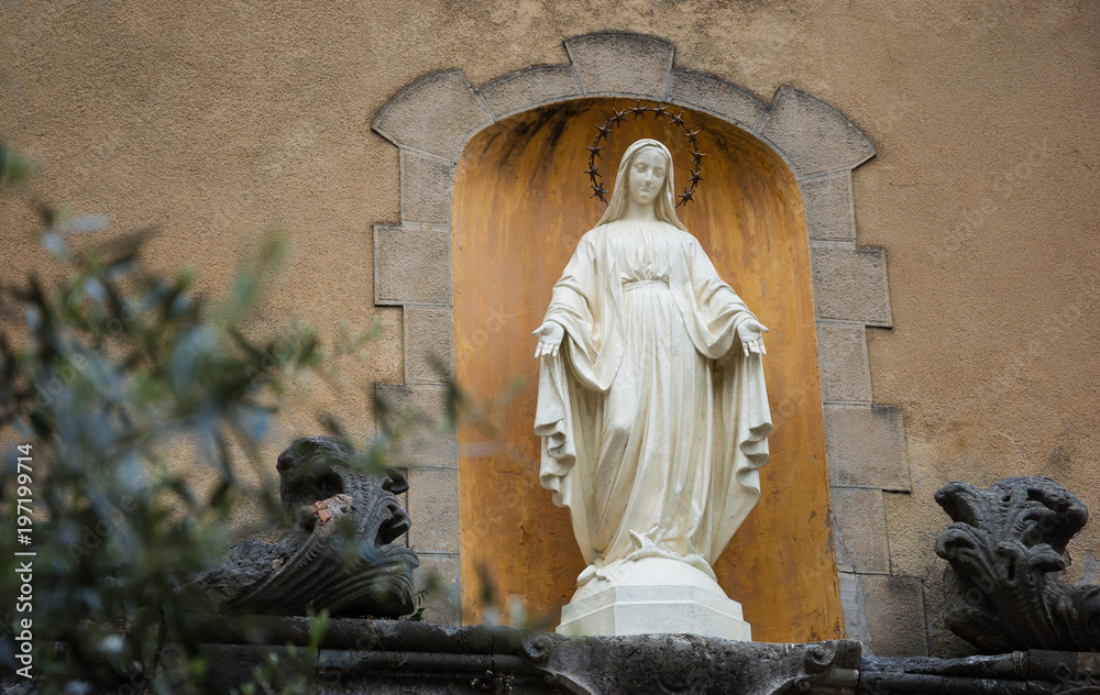 Φωτογραφία Statue of Madonna with stars nimbus on the front of Cathedral of Notre Dame of Assumption in Entraveaux (France) and olive tree at foreground
