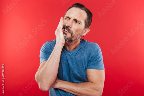 Portrait of adult bearded man suffering from toothache and touching jawbone with pain on face, isolated over red background