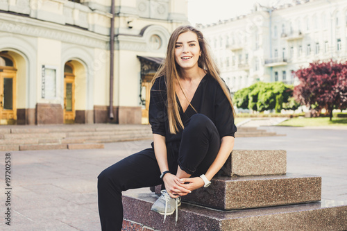 cute laughing model in black jumpsuit sitting at the street and trying to tie shoelaces photo