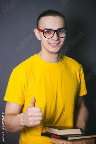 Fototapeta Naklejka Na Ścianę i Meble -  The guy in the yellow T-shirt and big glasses with books in their hands. Studio portrait on gray background. Student with books. photoshoot University