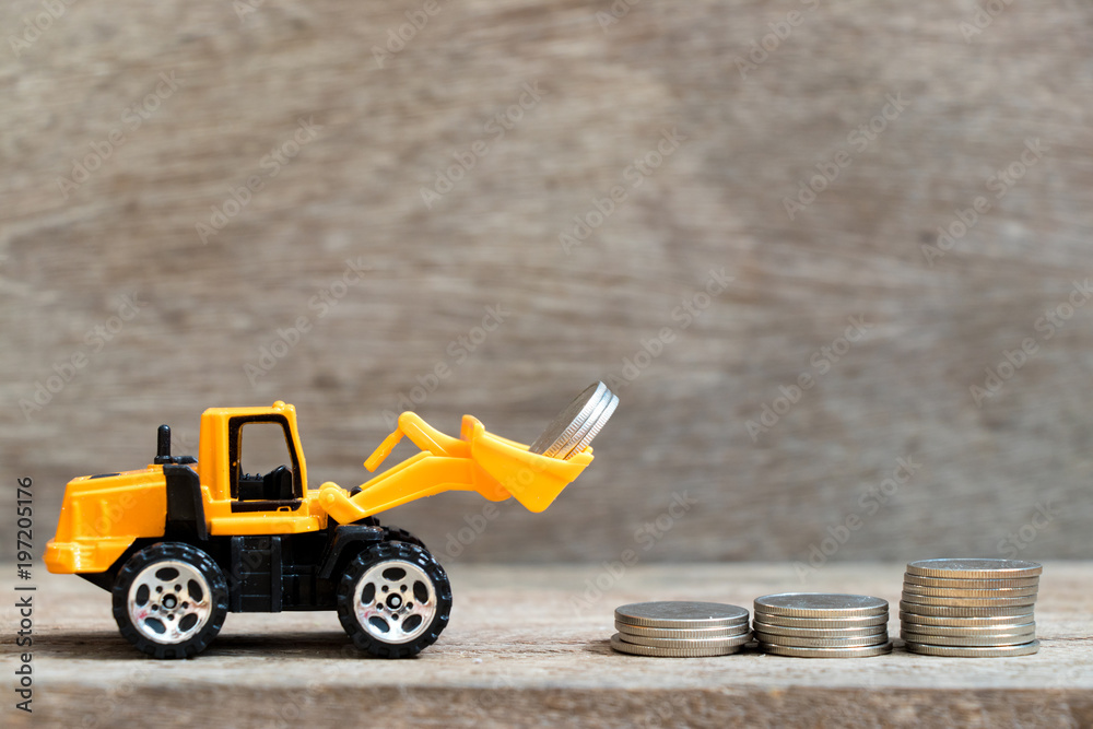 Fototapeta premium Toy bulldozer hold group of coin to fulfill up trend of stacked coin on wood background