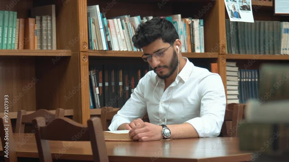 Good-looking asian man reading book, serious guy with black wavy hair wearing white headphones and glasses, sitting at the brown table next to the library bookshelves Good-looking asian man reading book, serious guy with black wavy hair wearing white headphones and glasses, sitting at the brown table next to the library bookshelves