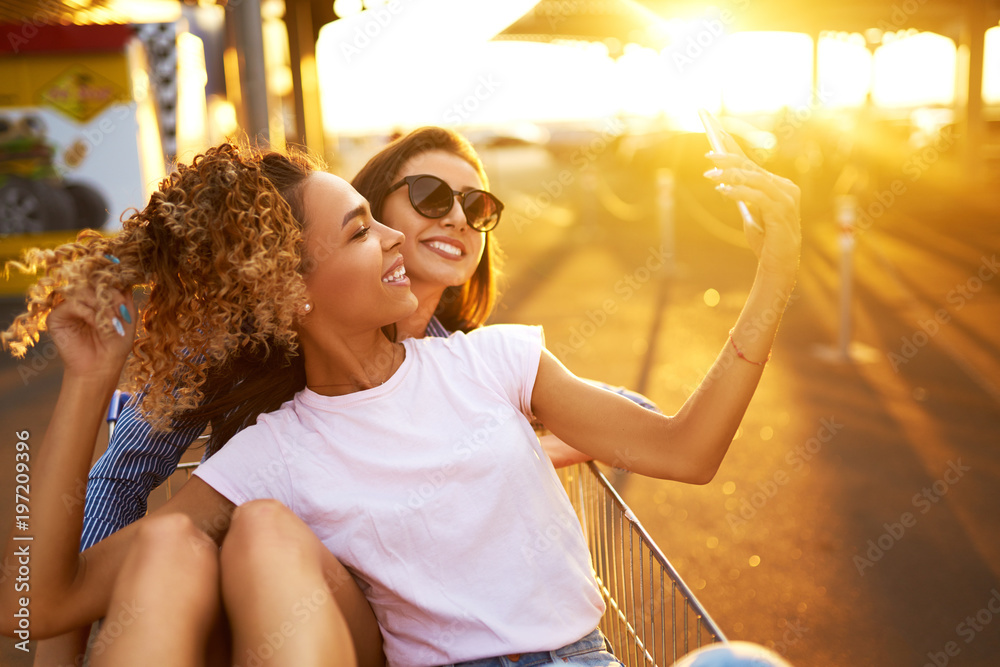 Fototapeta premium Selfie time! Two beautiful girls having fun on shopping trolleys. Multiethnic young people racing on shopping cart. Beautiful summer day with sunlight. Lifestyle concept.