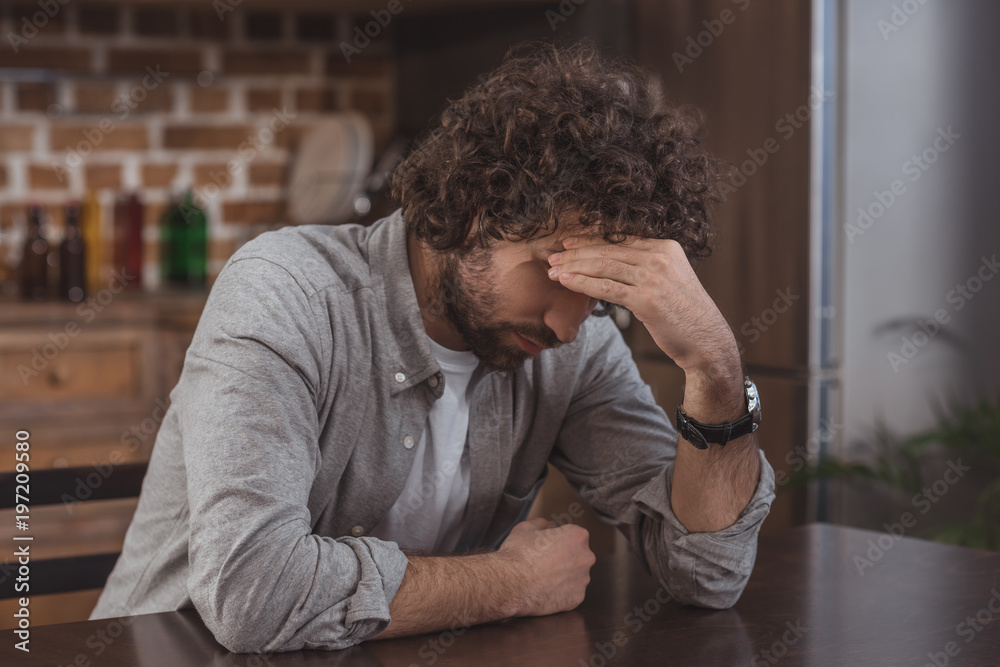 tired man sitting at table in kitchen and touching head