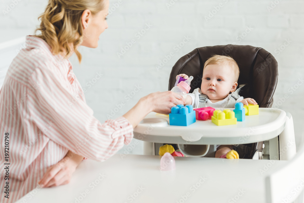 Mother feeding infant daughter sitting in baby chair with plastic blocks Mother feeding infant daughter sitting in baby chair with plastic blocks