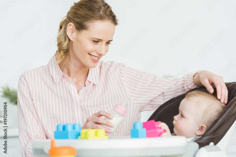 Mother with feeding bottle and infant daughter sitting in baby chair with plastic blocks