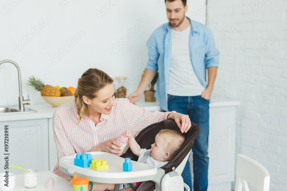 Mother giving feeding bottle to infant daughter in baby chair while father standing behind Mother giving feeding bottle to infant daughter in baby chair while father standing behind