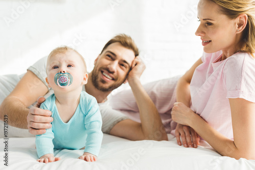 Smiling parents and infant daughter with baby dummy in bed