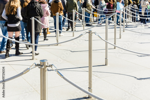 Stainless steel poles linked by grey ropes for queue control at the entrance of a tourist site, with drop shadows on a light ground and blurry people queueing up in the background.