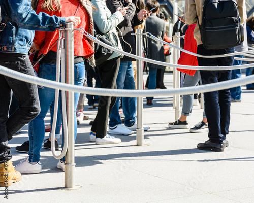 People standing in line between stainless steel queue poles with grey ropes, at the entrance of a tourist site, leaning on the poles or watching their smartphones. photo
