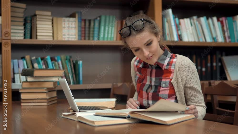 Smiling caucasian girl studying in library, doing research with pleasure with use of the internet and books, wearing checked casual shirt and glasses on the head, concept of positive attitude to Smiling caucasian girl studying in library, doing research with pleasure with use of the internet and books, wearing checked casual shirt and glasses on the head, concept of positive attitude to