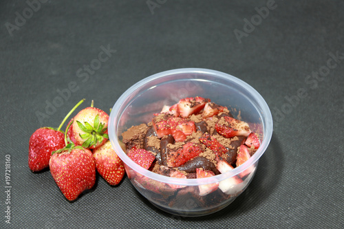 Home made cake and chocolate lava, topping by Strawberries cut into pieces with cocoa powder in the round plastic cup. And Strawberry on the black floor.