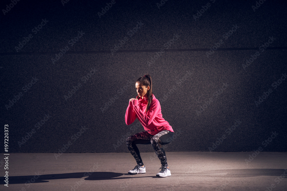 Portrait view of young sexy attractive healthy fitness sporty active slim girl with sportswear crouching and doing squats while holding hands together in front of the grey wall at night on the street.