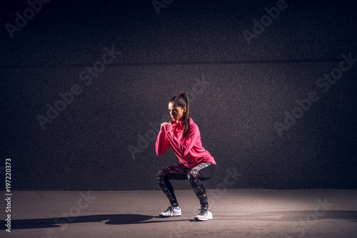 Portrait view of young sexy attractive healthy fitness sporty active slim girl with sportswear crouching and doing squats while holding hands together in front of the grey wall at night on the street.