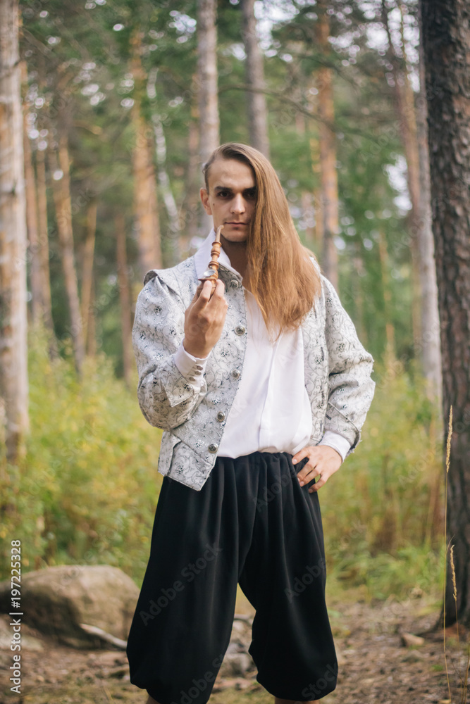 Sexy long-haired man with a pipe in his hands, in the woods. In a beautiful, ancient jacket with patterns and a white shirt. Fantasy. Sexy long-haired man with a pipe in his hands, in the woods. In a beautiful, ancient jacket with patterns and a white shirt. Fantasy.