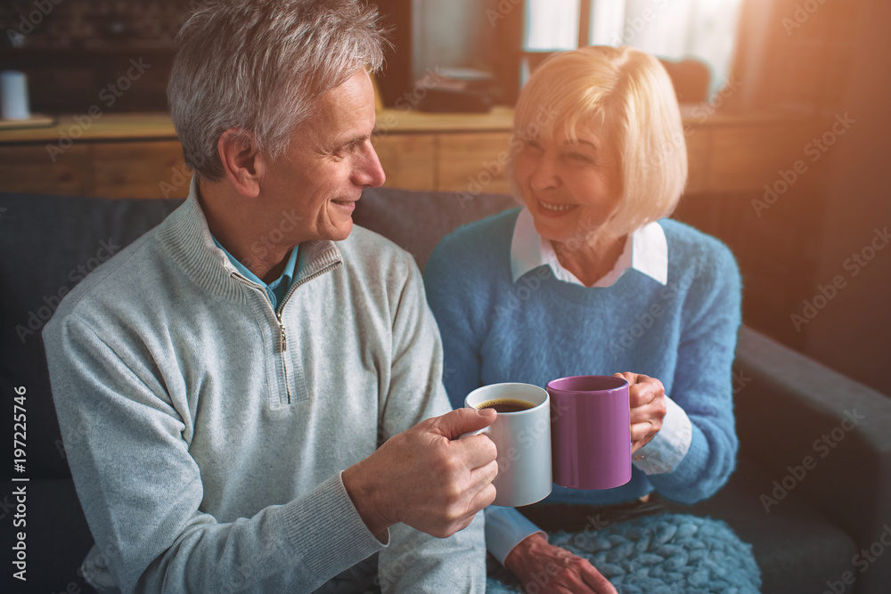 Husband and wife are sitting together and drinking tea from cups. They are looking to each other and smiling. Husband and wife are sitting together and drinking tea from cups. They are looking to each other and smiling.