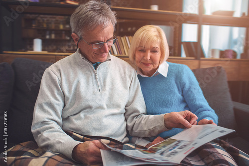 Beautiful old couple are sitting on the couch together and reading a big newspaper. Man is using reading glasses. They look concentrated and consious. photo