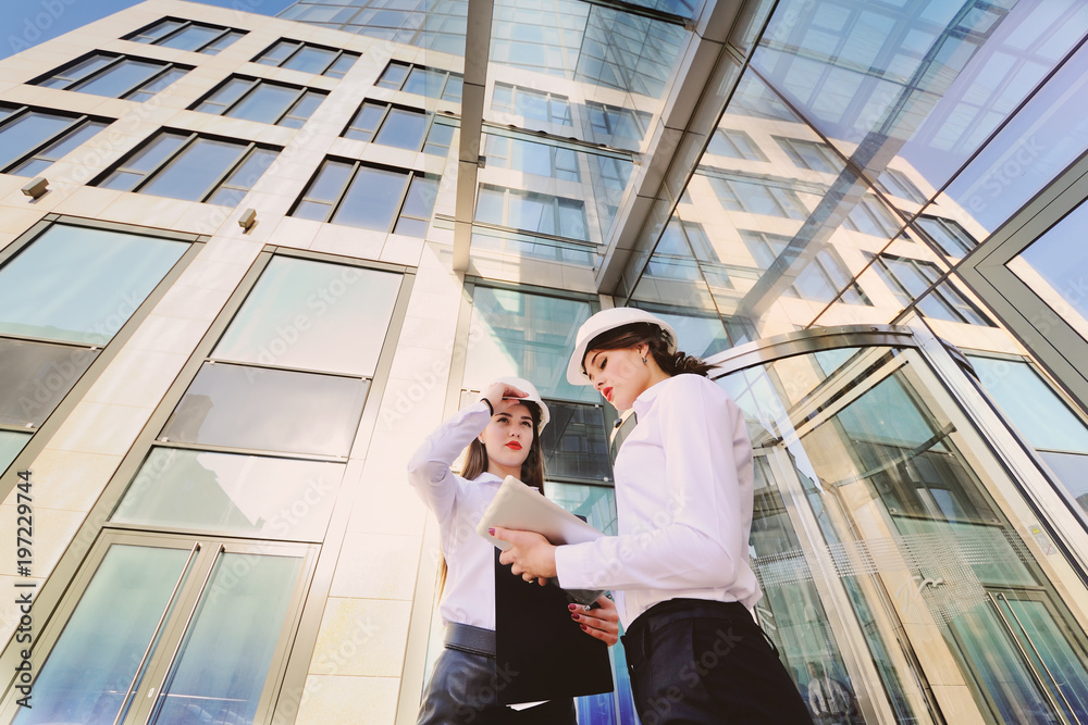 Two young pretty business women industrial engineers in construction helmets with a tablet in hands on a glass building background Two young pretty business women industrial engineers in construction helmets with a tablet in hands on a glass building background