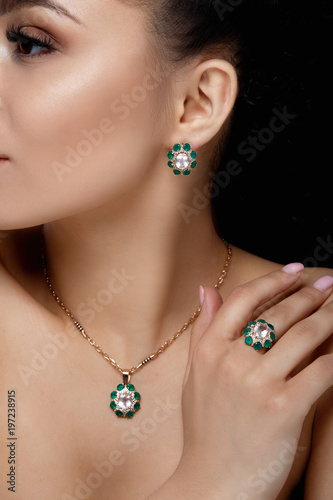 Charming model with dark hair shows rich golden earrings, necklace, and ring with green precious stones