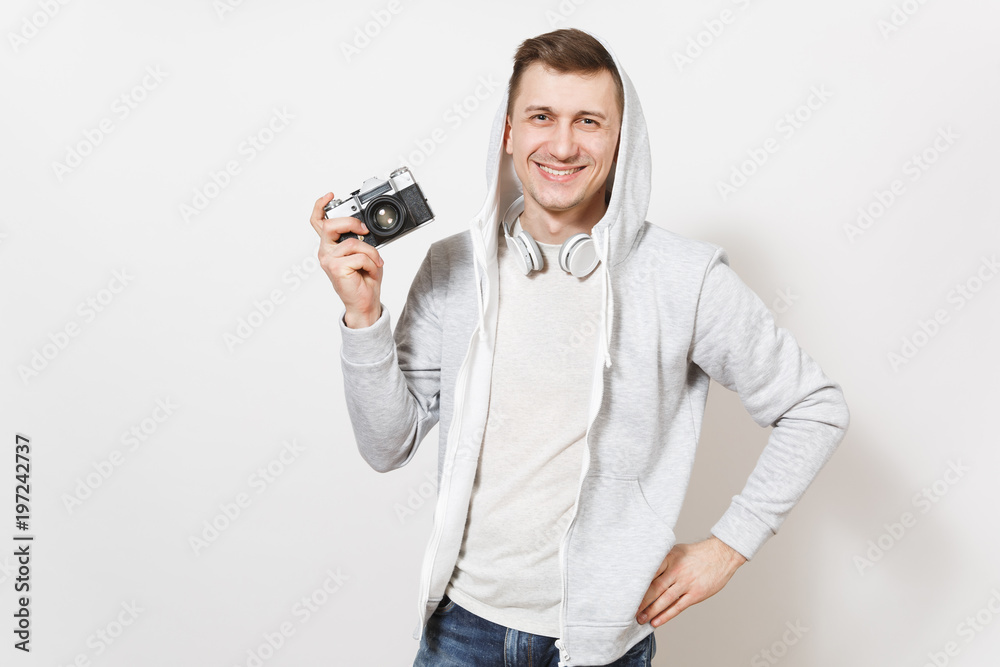 Young handsome smiling male student in t-shirt and light sweatshirt with hood with headphones holds retro camera and holds hand on waist isolated on white background. Concept of photography, hobby Young handsome smiling male student in t-shirt and light sweatshirt with hood with headphones holds retro camera and holds hand on waist isolated on white background. Concept of photography, hobby
