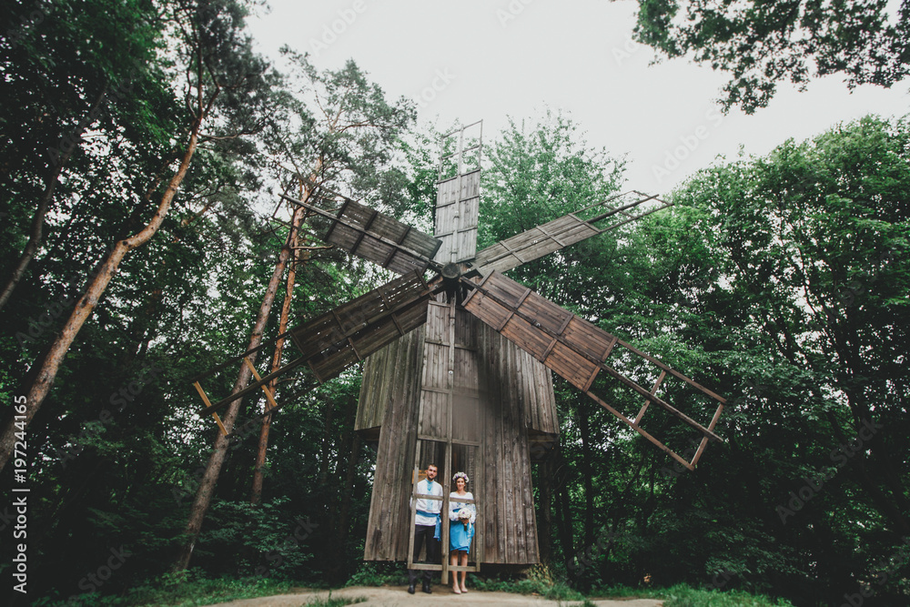 Traditional wedding couple in national dress is standing near old wooden mill. Romantic photo of groom and bride in love story. Beautiful flowers in ukrainian bride's crown. Vintage mill photo. Traditional wedding couple in national dress is standing near old wooden mill. Romantic photo of groom and bride in love story. Beautiful flowers in ukrainian bride's crown. Vintage mill photo.