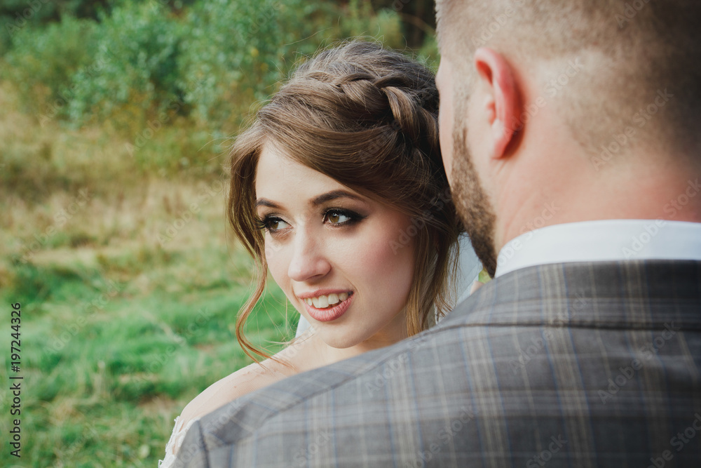 Beautiful wedding couple in the forest. The bride in tulle veil turns her head to the side near the bearded groom in checkered suit. Stylish and rustic love story. Amazing bridal portrait. Beautiful wedding couple in the forest. The bride in tulle veil turns her head to the side near the bearded groom in checkered suit. Stylish and rustic love story. Amazing bridal portrait.