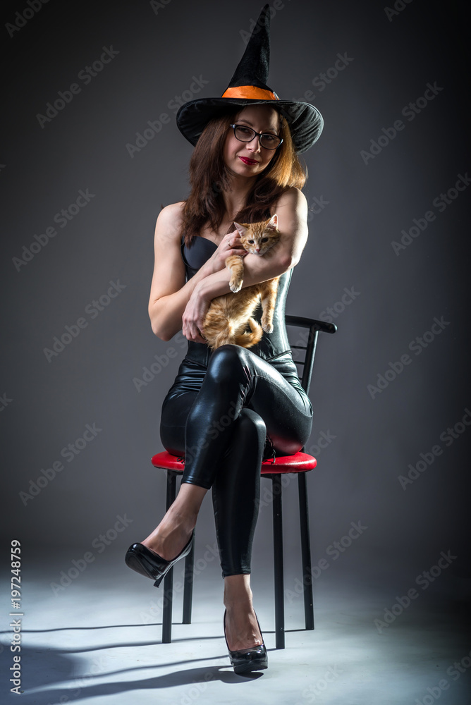 Woman in witch costume sitting on a chair and holding a kitten, background Halloween.