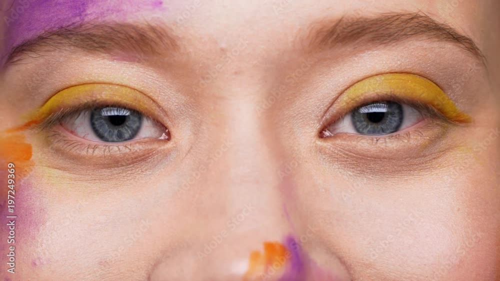 Extreme closeup portrait of european woman 20s with colorful painting on face, macro view of pleased eyes looking straight, and smiling in joy