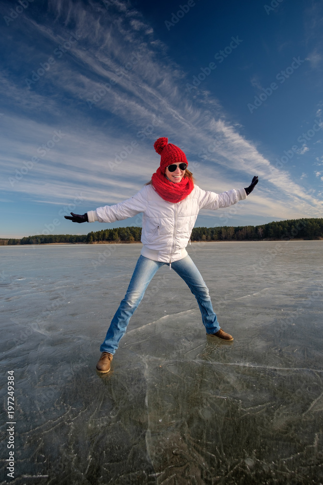 Girl in sunglasse, white jacket, red cap and red scarf walking and having fun on ice on the frozen lake in winter