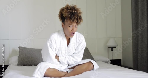 Young ethnic girl in white bathrobe sitting with eyes closed on bed and grimacing from pain in stomach. photo