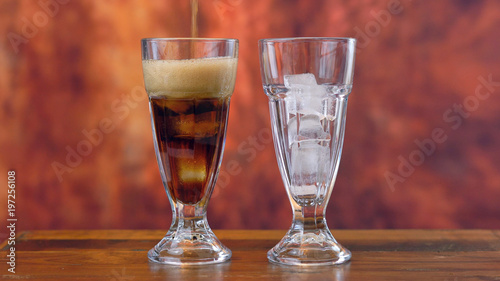 Pouring cola soft drink on ice in tall cafe glasses against a rustic wood background.